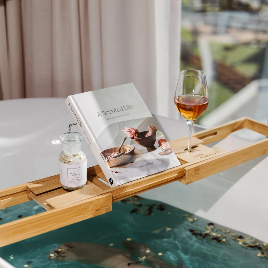 Wooden bath caddy resting on top of bath with a glass of wine, book and jar of bath salts on top