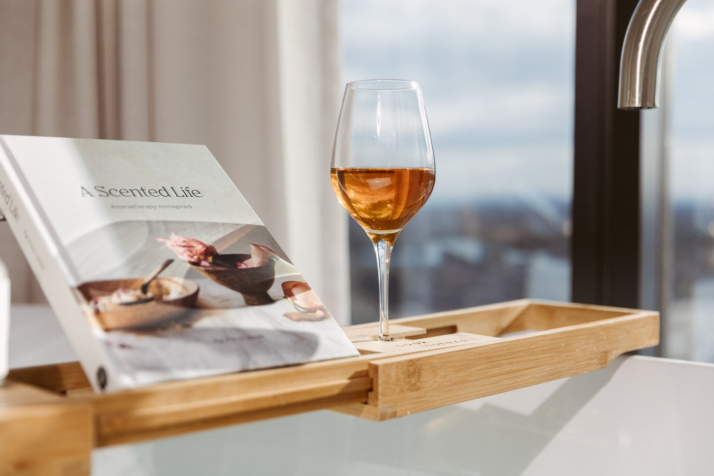 A bath tub with a bamboo caddy across it. On the shelf is a glass of rose wine and a book. The book is titled "A scented life"