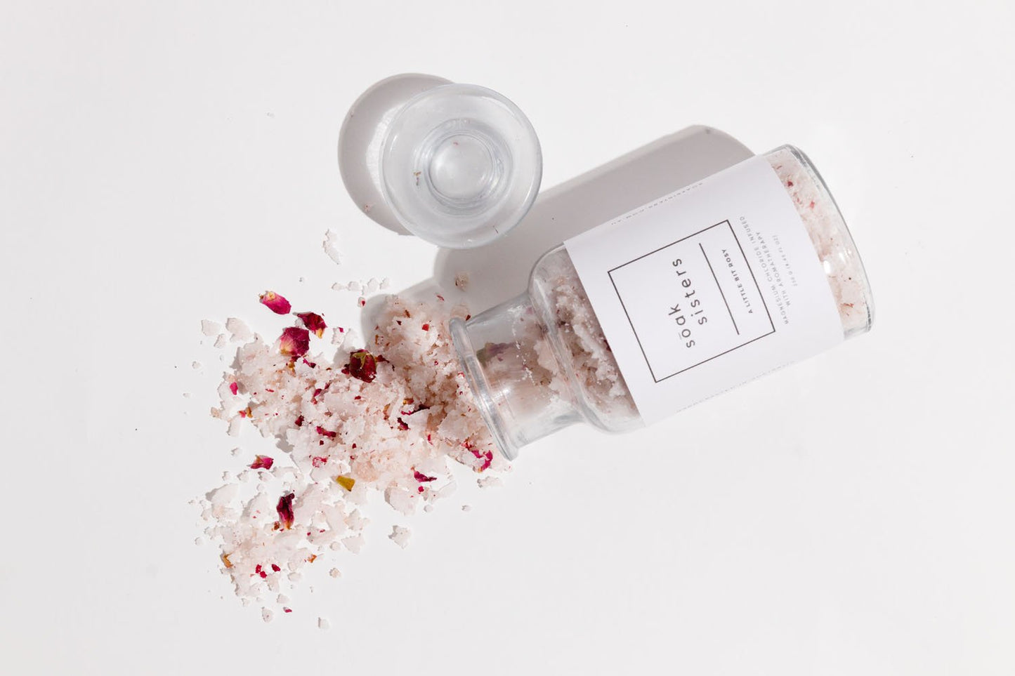 Jar of A Little Bit Rosy on white surface with bath salts spilling out