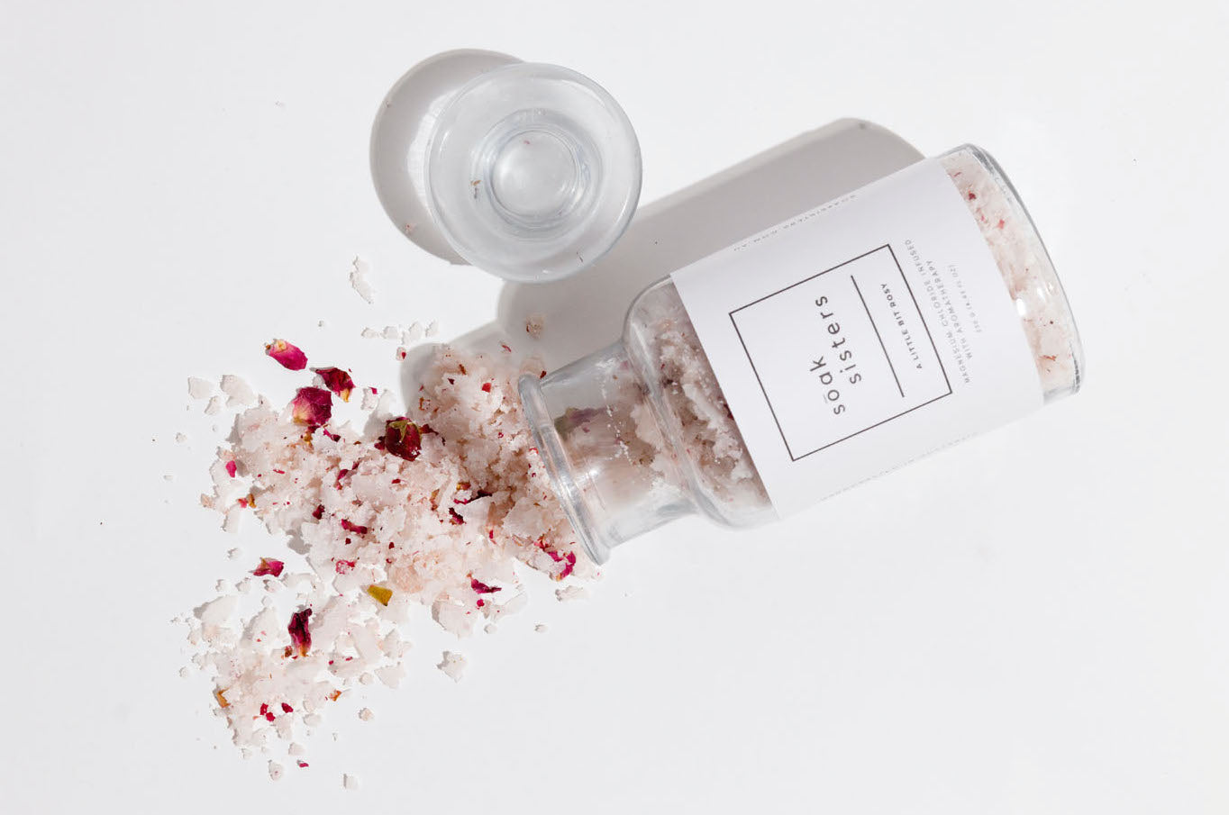 A bottle of bath salts laying on its side with the lid off. Some of the bath salts have spilled out of the bottle. The label on the bottle says "soak sisters"