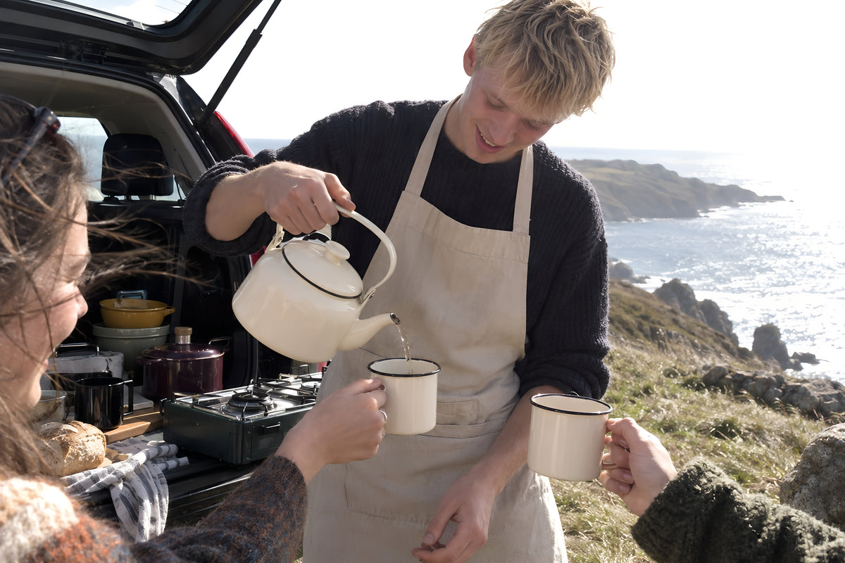 The cream KILN Kettle on a camping stove at the open boot of a car on a windy headland, two mugs held ready. Daytime, hard sun, no candles lit.