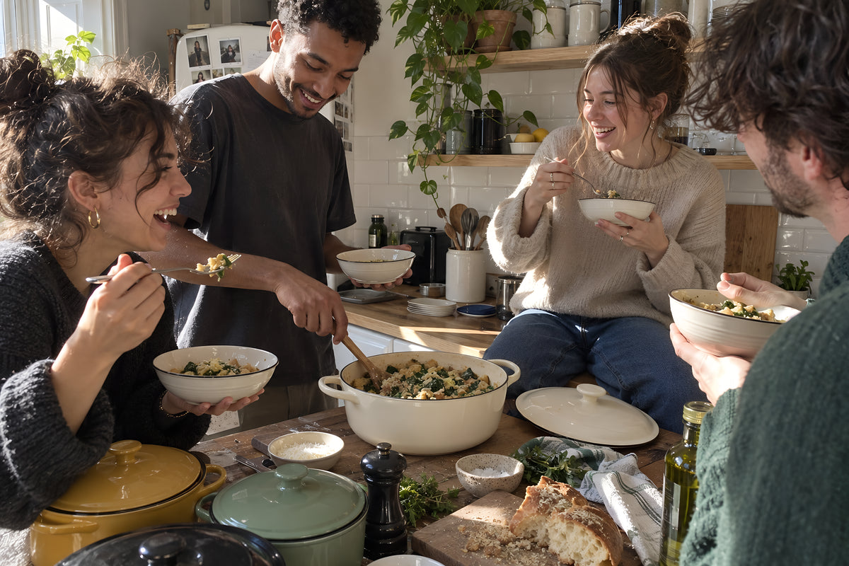 Four housemates eating straight out of the cream KILN Casserole standing round a tiny kitchen, bowls in hands, one sitting on the worktop. Daytime, hard sun, no candles lit.